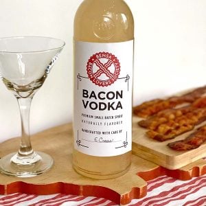 bottle of bacon vodka on a wooden cutting board, an empty martini glass, and four strips of crisply cooked bacon