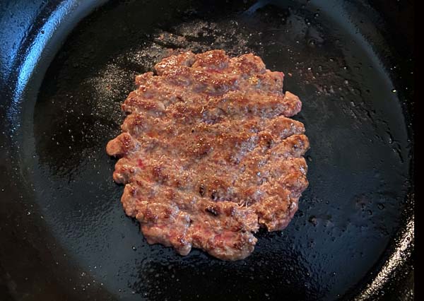 Hamburger patty cooking in a cast iron skillet.