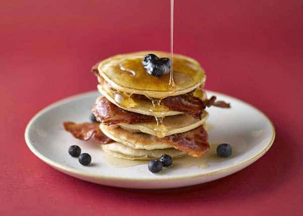 syrup being poured over bacon fat pancakes, which are layered with strips of bacon and blueberries