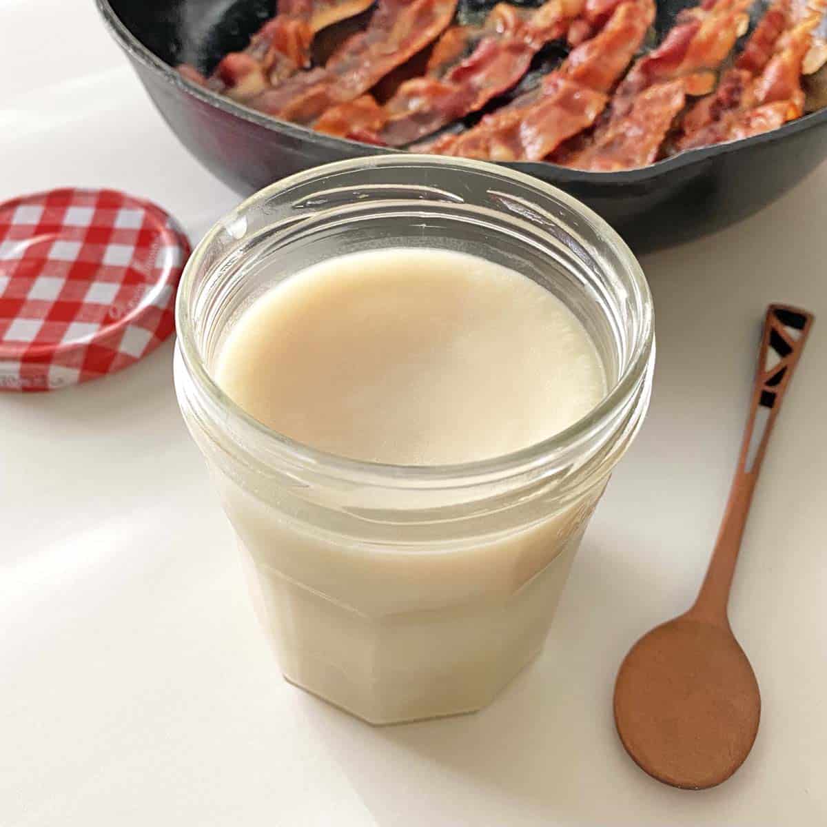 A jar filled with refrigerated bacon fat, in front of a frying pan with six strips of golden brown bacon. 