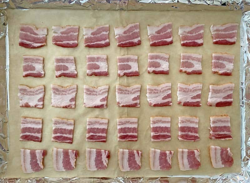 Squares of bacon arranged on a baking sheet lined with foil and parchment paper