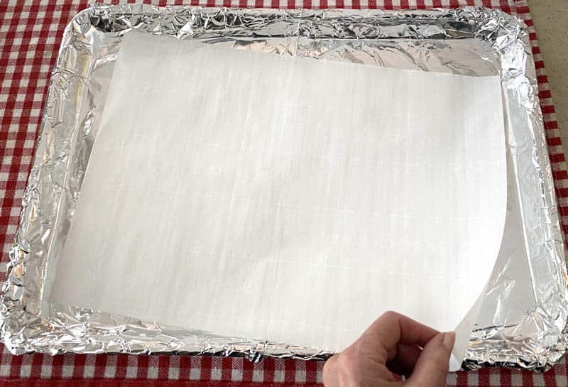 A hand laying a sheet of white parchment paper over a baking sheet lined with foil.
