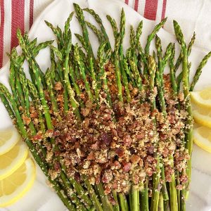 Asparagus spears on a white platter, topped with crumbled bacon, bread crumbs and Parmesan cheese, garnished with six lemon wedges.