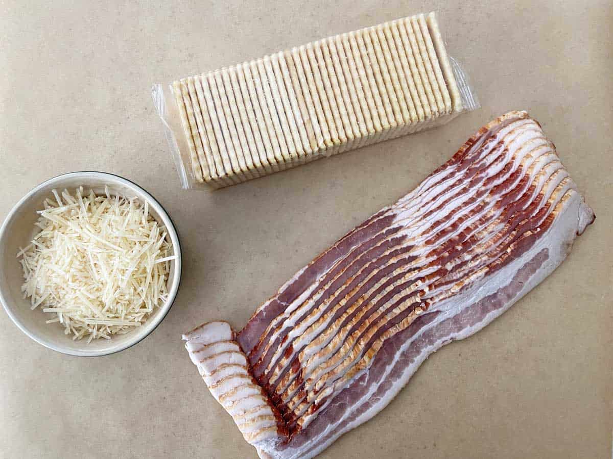 A pound of bacon, a sleeve of 36 club crackers, and a white bowl with shredded Parmesan cheese