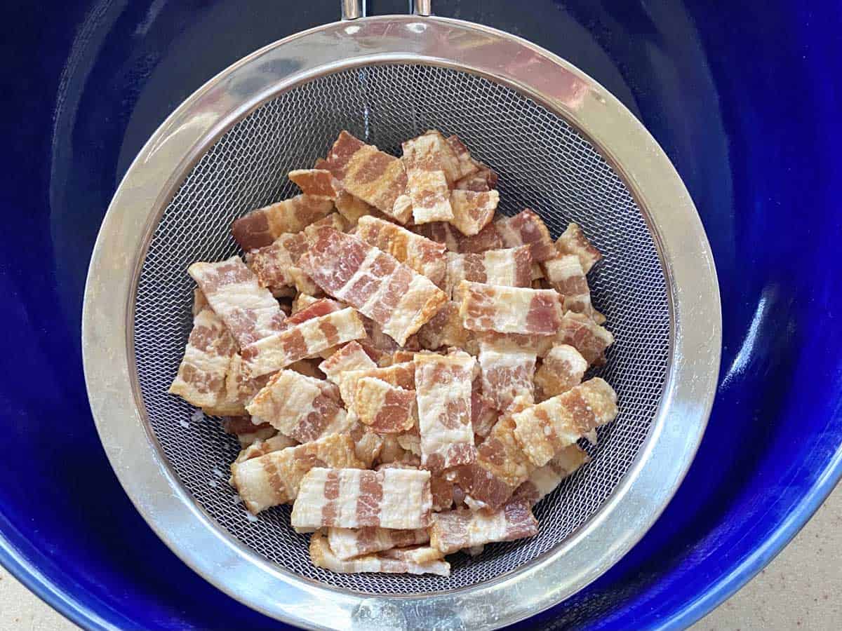 A strainer over a blue bowl with the bacon pieces (they look weird and pale) being strained out.