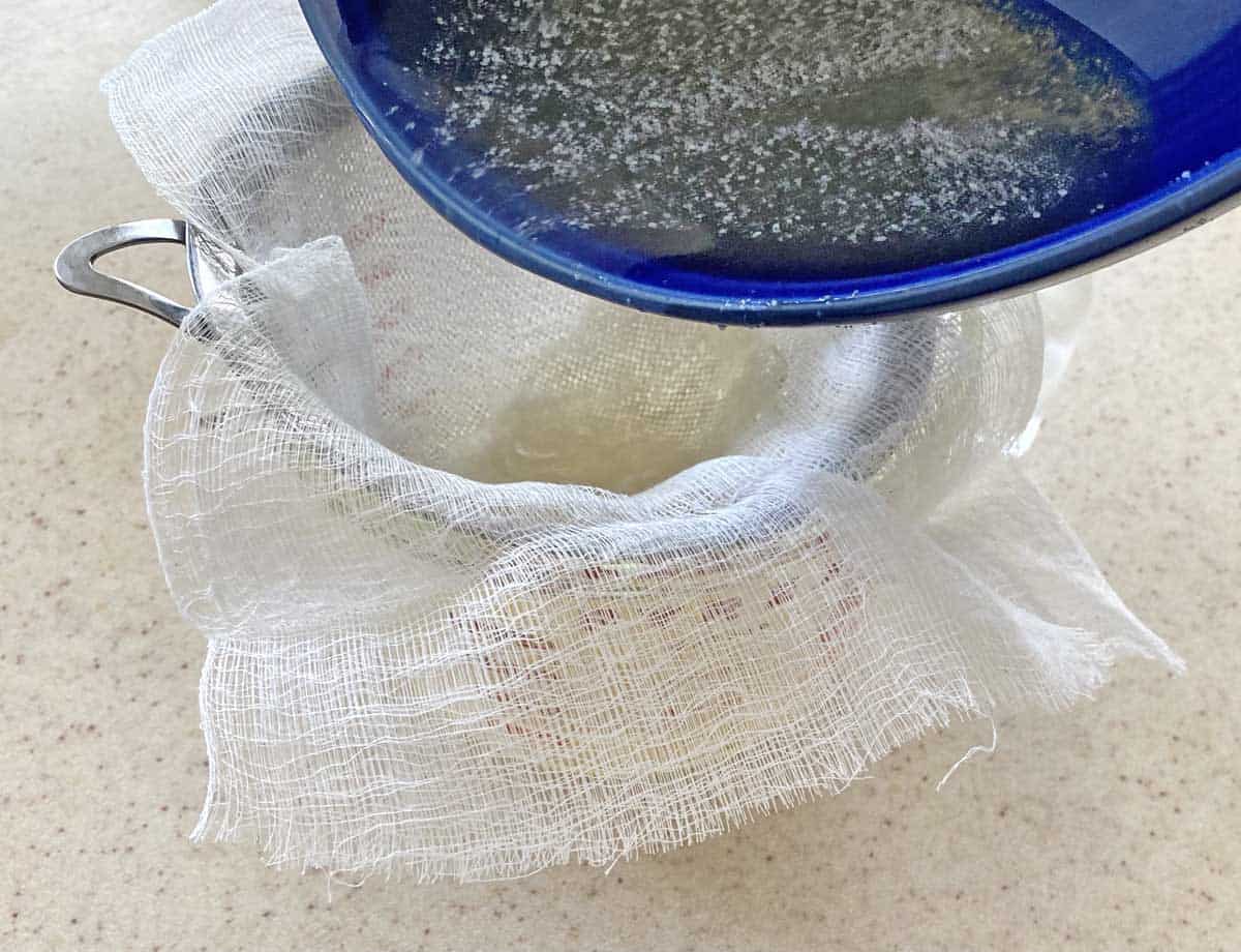 A blue bowl filled with the bacon vodka mixture is being poured through a cheesecloth lined strainer into a large measuring cup. 