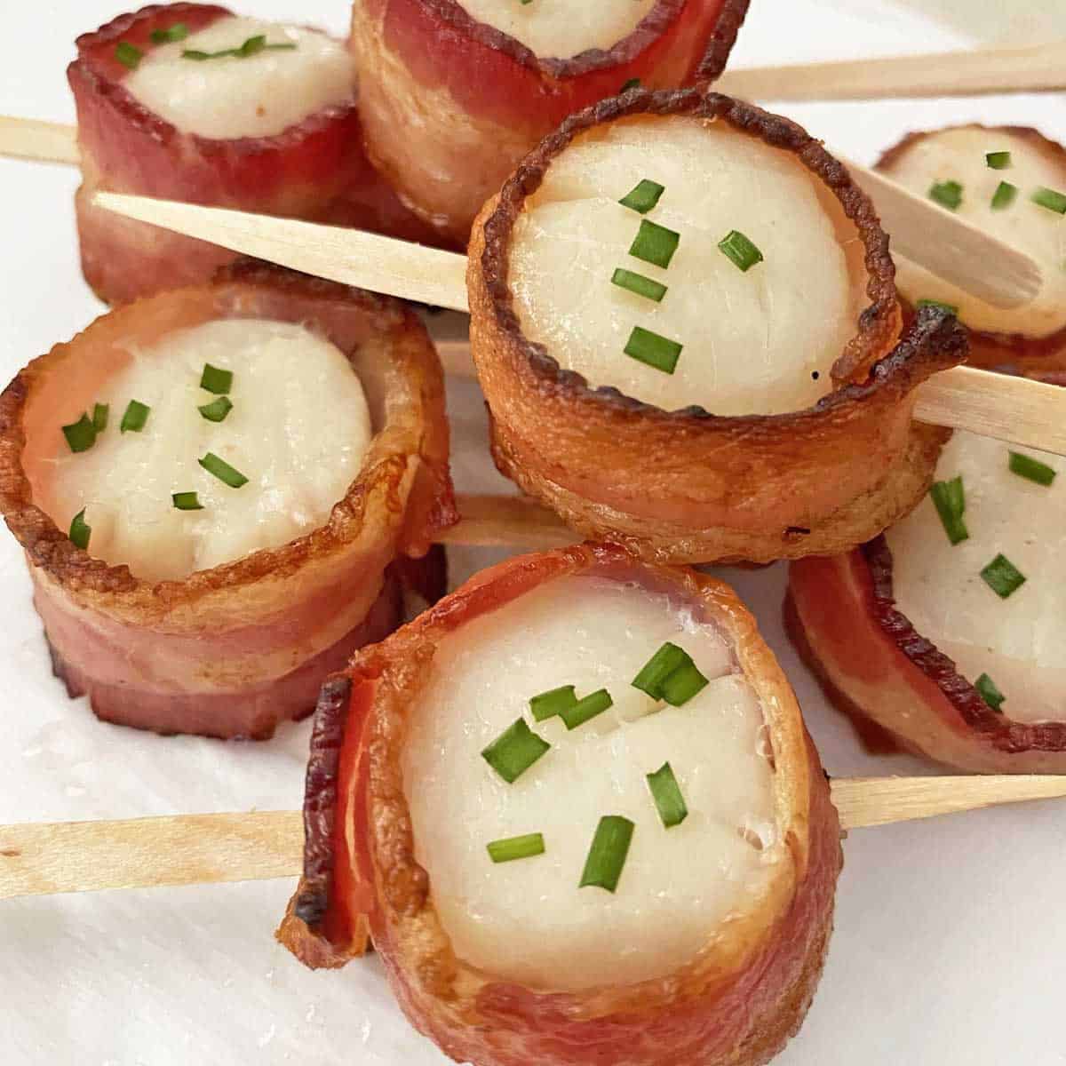 A pile of bacon wrapped scallops garnished with chives on a white background.