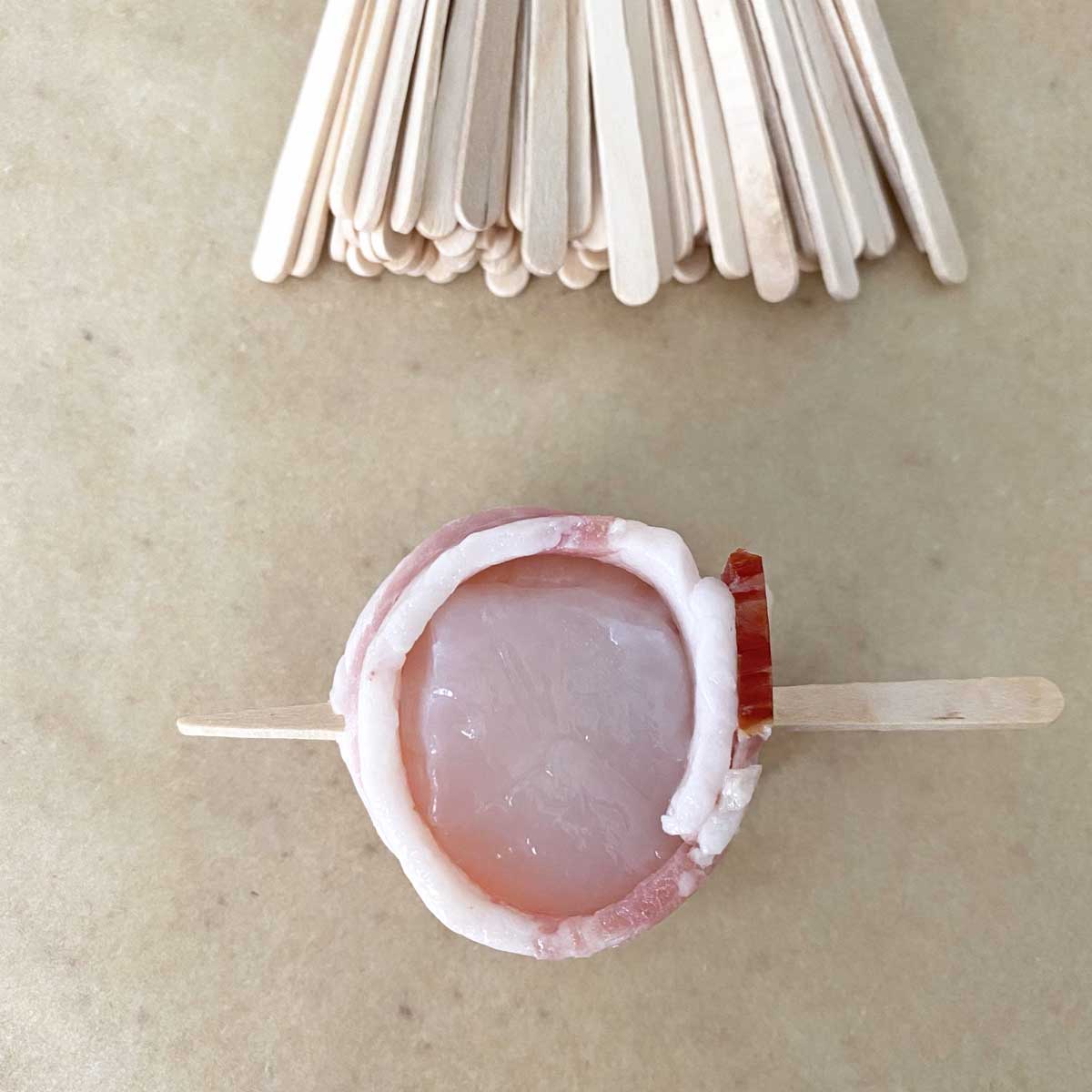 A single uncooked bacon wrapped scallop and a pile of wooden picks