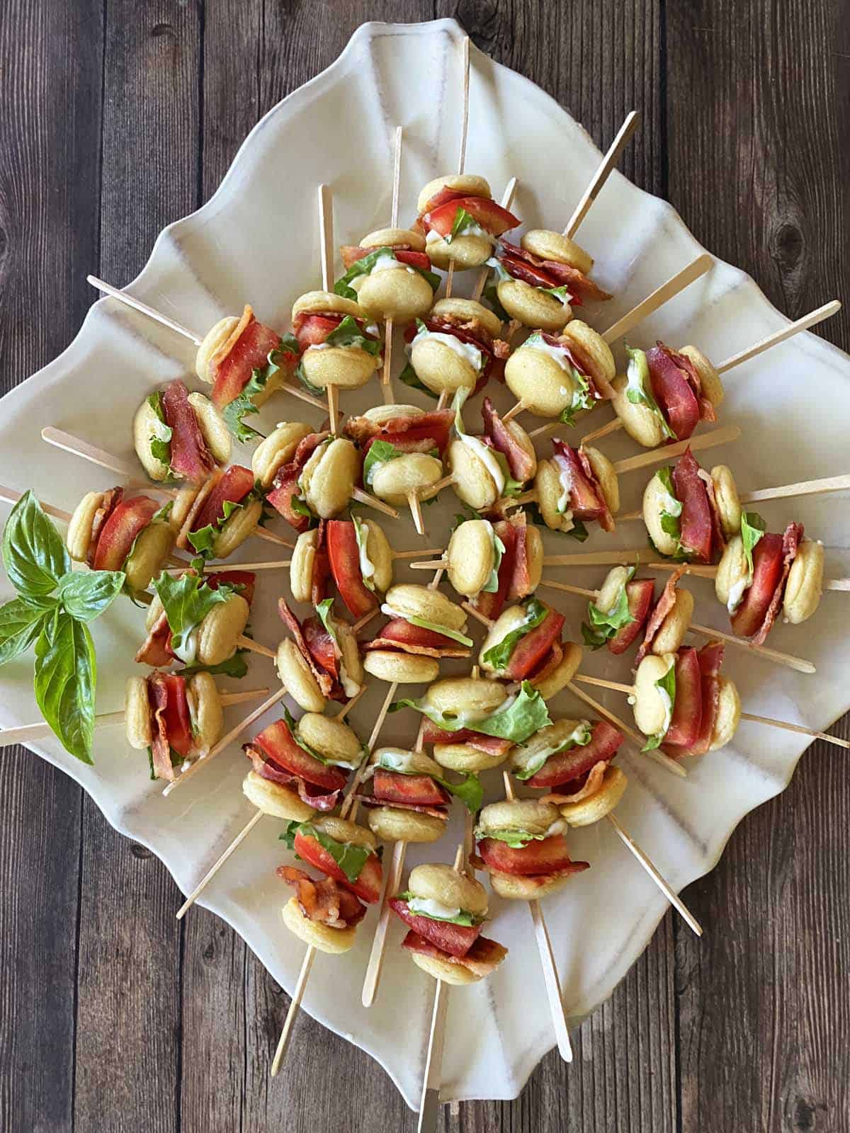 Mini BLT sliders on skewers arranged on a white plate against a wood background