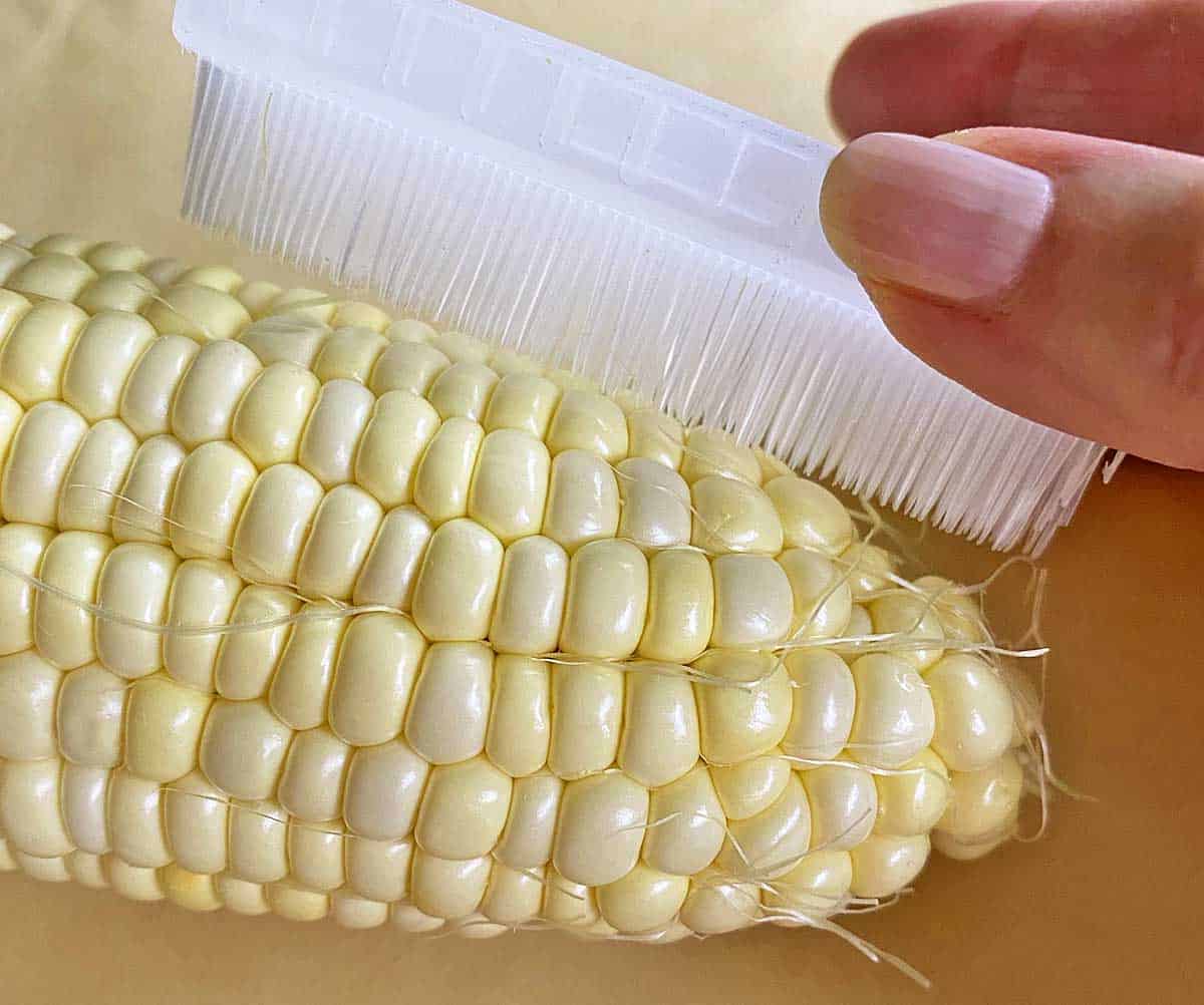 Brushing an ear of uncooked corn on the cob with a corn silk brush