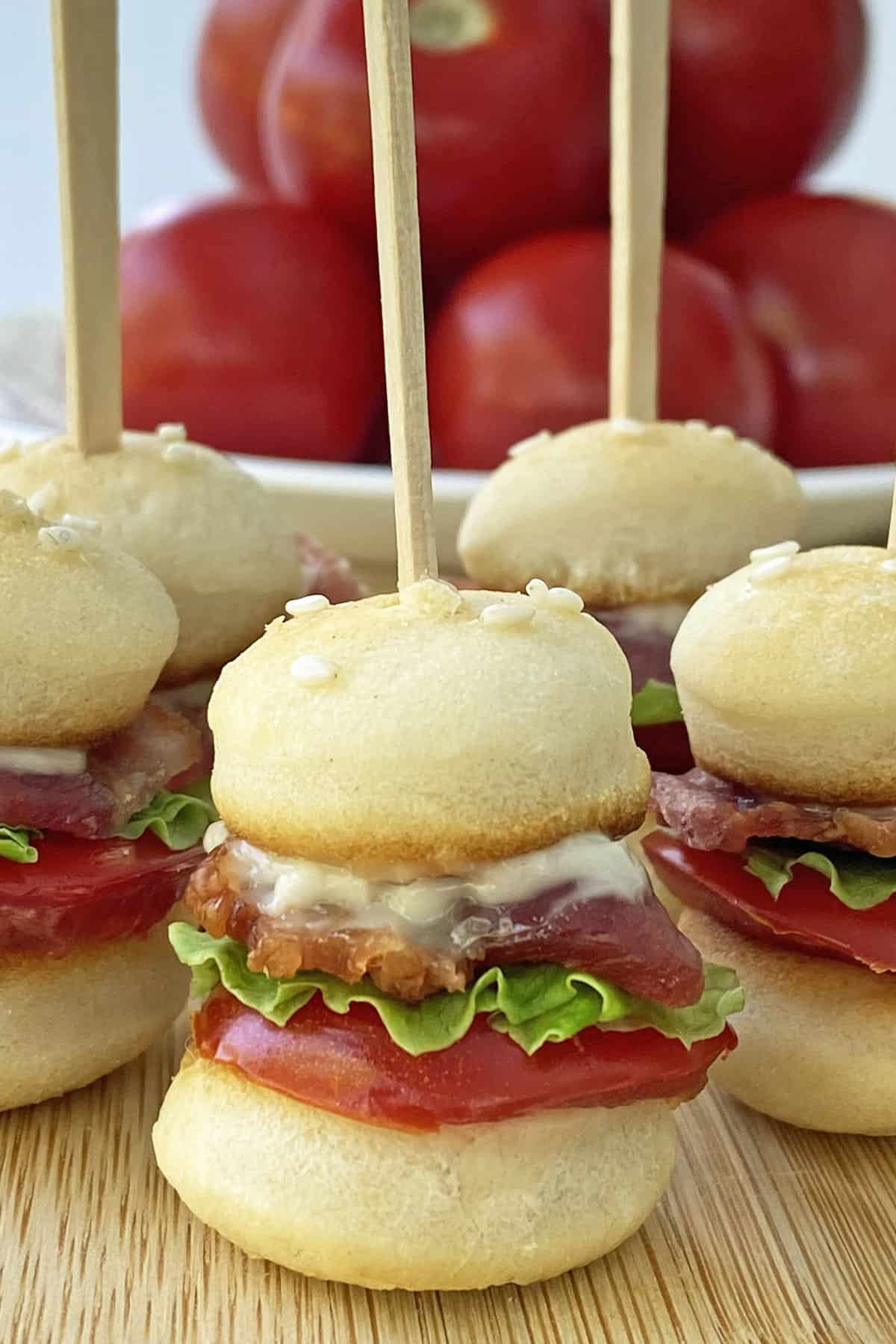 Close up of mini BLT sliders on toothpicks on a wooden cutting board with a bowl of tomatoes in the background. 