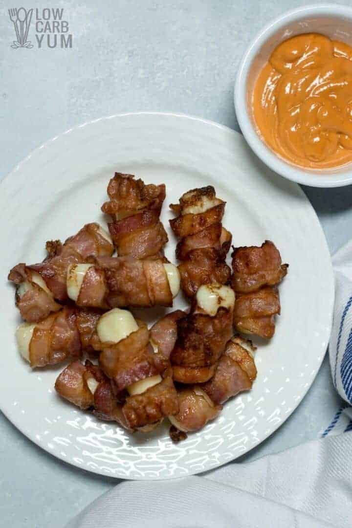 A pile of bacon wrapped cheese sticks on a white platter with dipping sauce on the right
