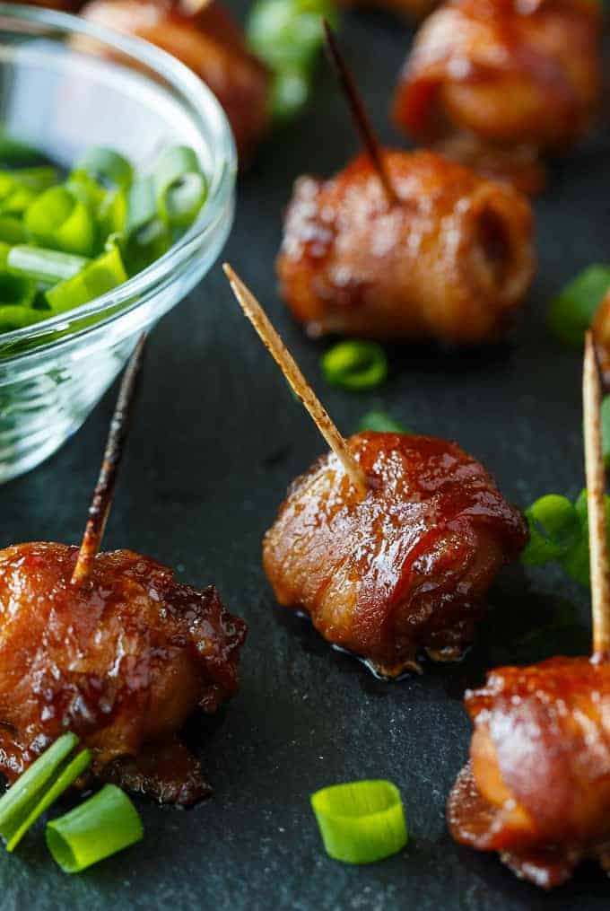 7 pieces of bacon wrapped water chestnuts on a black background with dipping sauce on the left