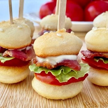 Closeup of 5 mini BLT sliders on skewers resting on a bamboo cutting board, with a bowl of tomatoes in the background