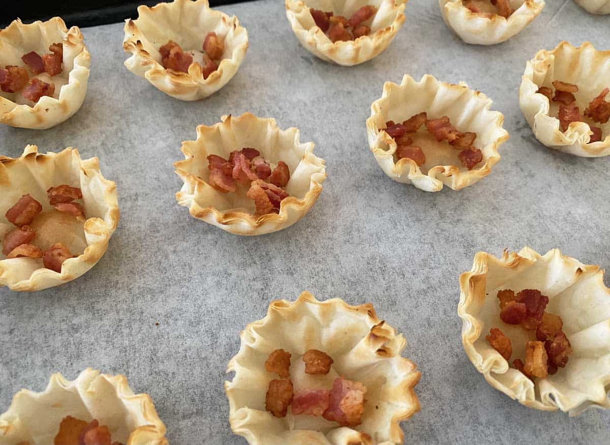 A dozen phyllo shells filled with bacon bits on a parchment paper-lined baking sheet.