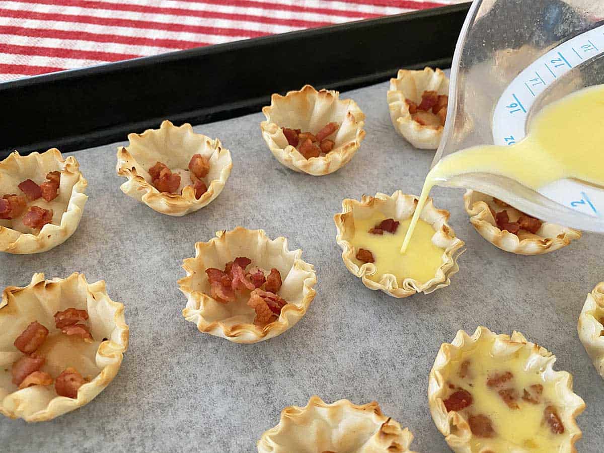 A measuring cup filled with egg mixture being poured into a phyllo cup on a baking sheet with a dozen more cups.