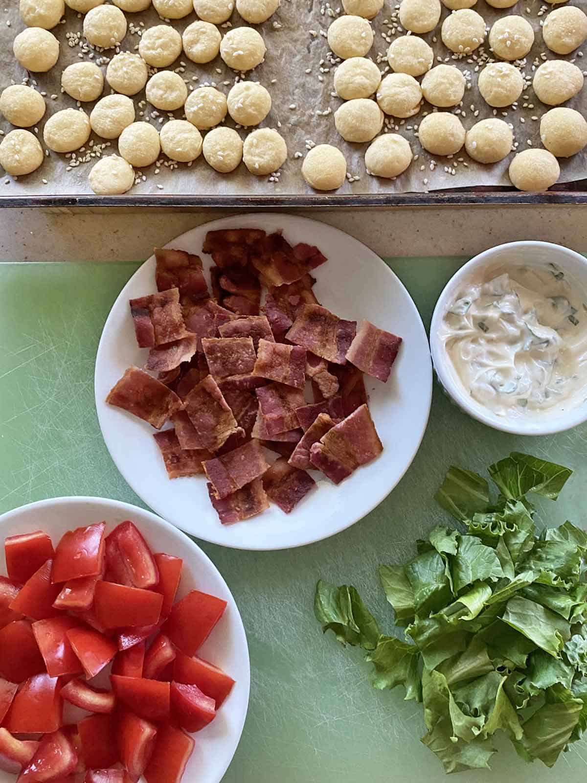 Baby buns on a tray, cooked bacon squares on a white dish, basil mayonnaise, chopped tomatoes, chopped lettuce ready to assemble sliders.