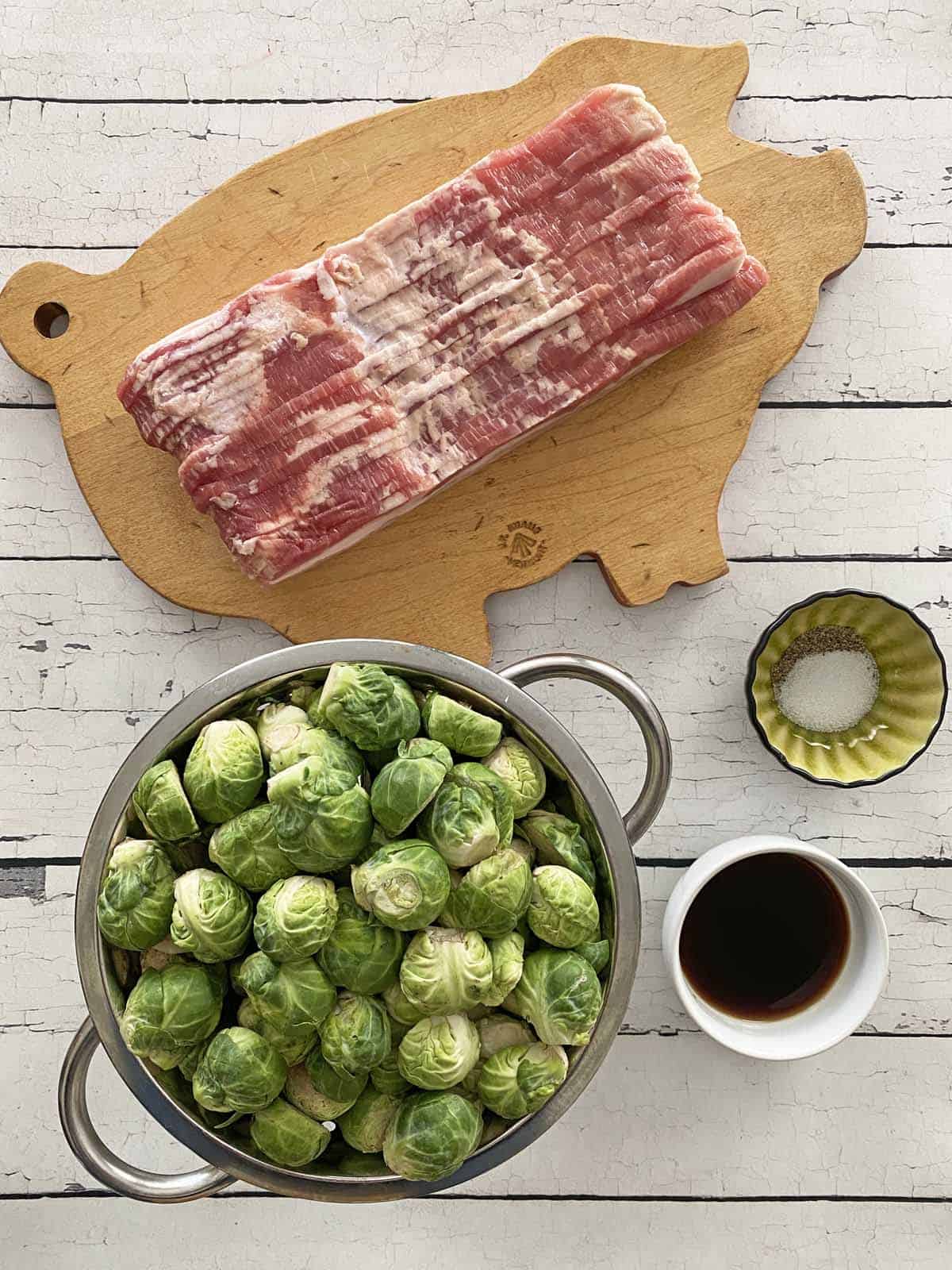 A wood cutting board with a pound of bacon, a collander full of Brussels sprouts, a dish of balsamic vinegar and a dish of salt and pepper