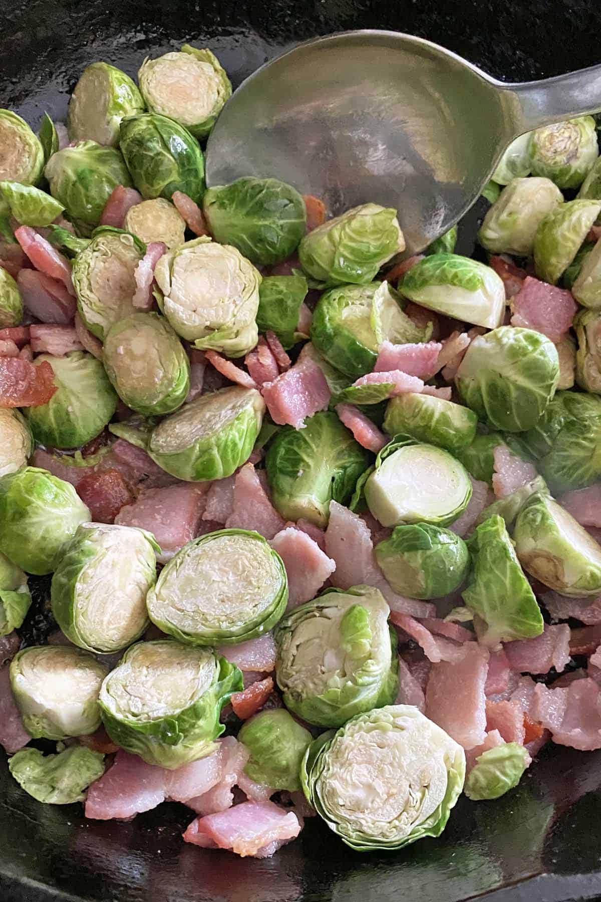 Partially cooked steamed Brussel sprouts with partially cooked bacon pieces in a skillet with a spoon