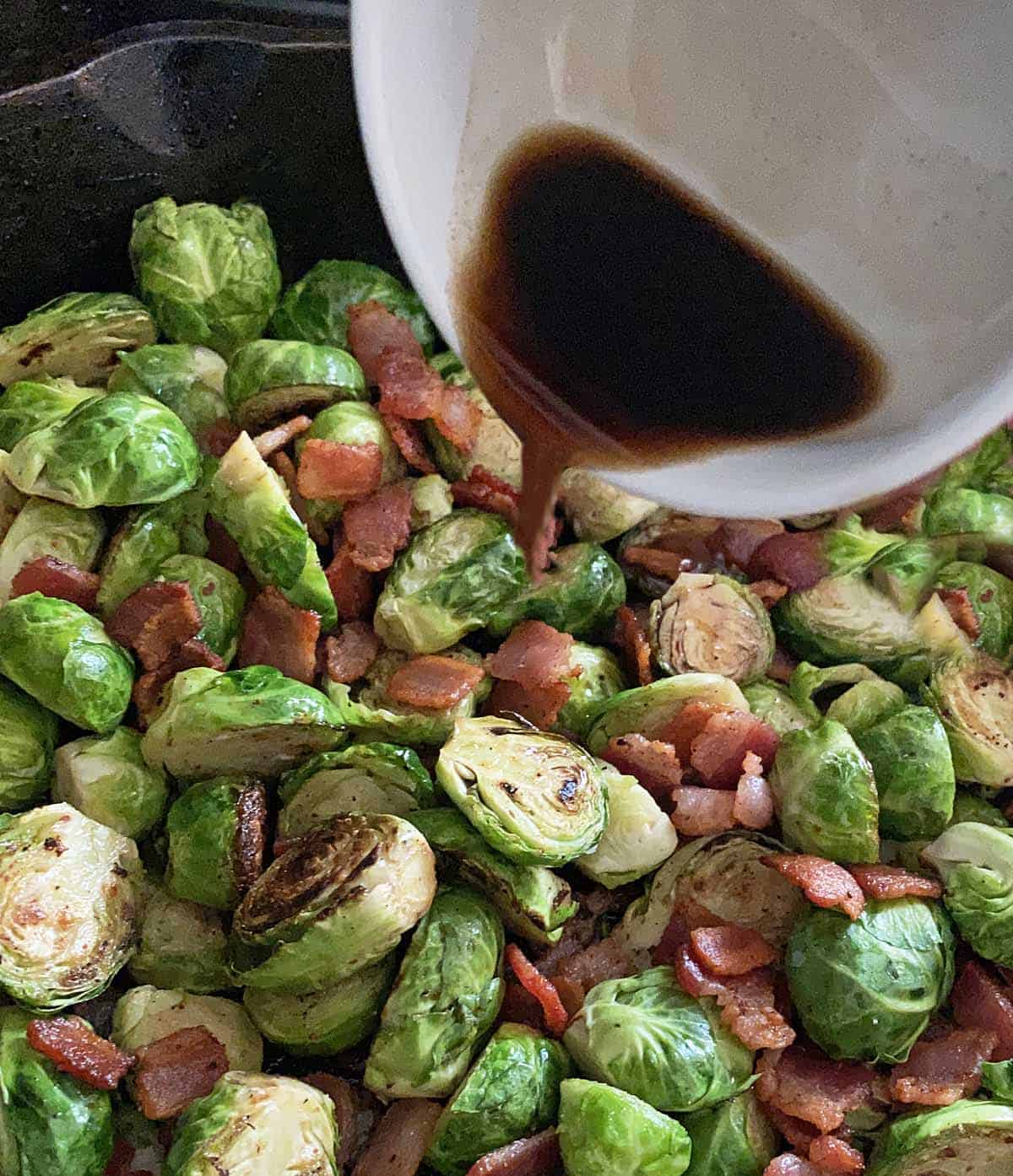 A close up of a small white dish of balsamic vinegar being drizzled over Brussel sprouts with bacon