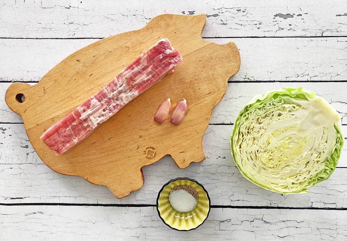 8 slices of bacon and 2 garlic cloves on a pig-shaped wooden cutting board, a cabbage half and a small dish with salt and pepper.