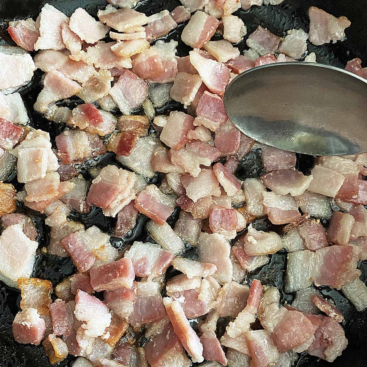 Close up of partially cooked chopped bacon in a skillet with a spoon stirring it