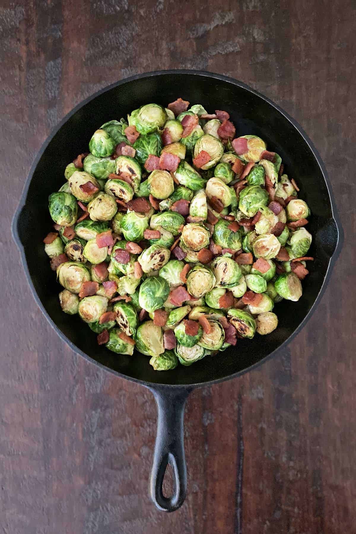 A cast iron skilled filled with cooked Brussel sprouts and bacon, set on a dark brown wooden table.