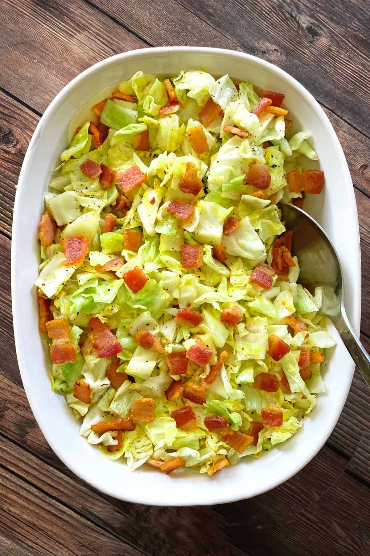 White oval serving dish filled with fried cabbage with bacon and garlic and a silver serving spoon.