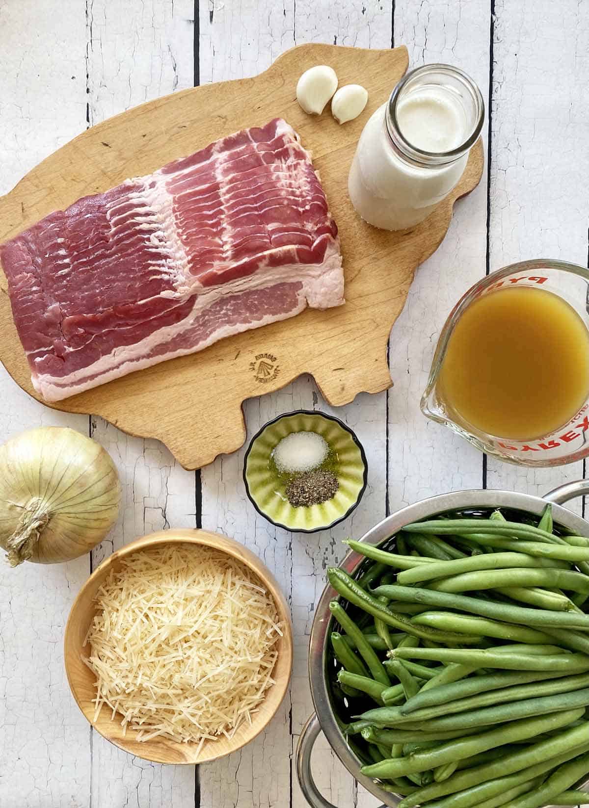 A wooden cutting board on a white rustic background with bacon, cream, 2 garlic cloves, chicken stock, green beans, Parmesan cheese, and onion and small dish with salt and pepper.