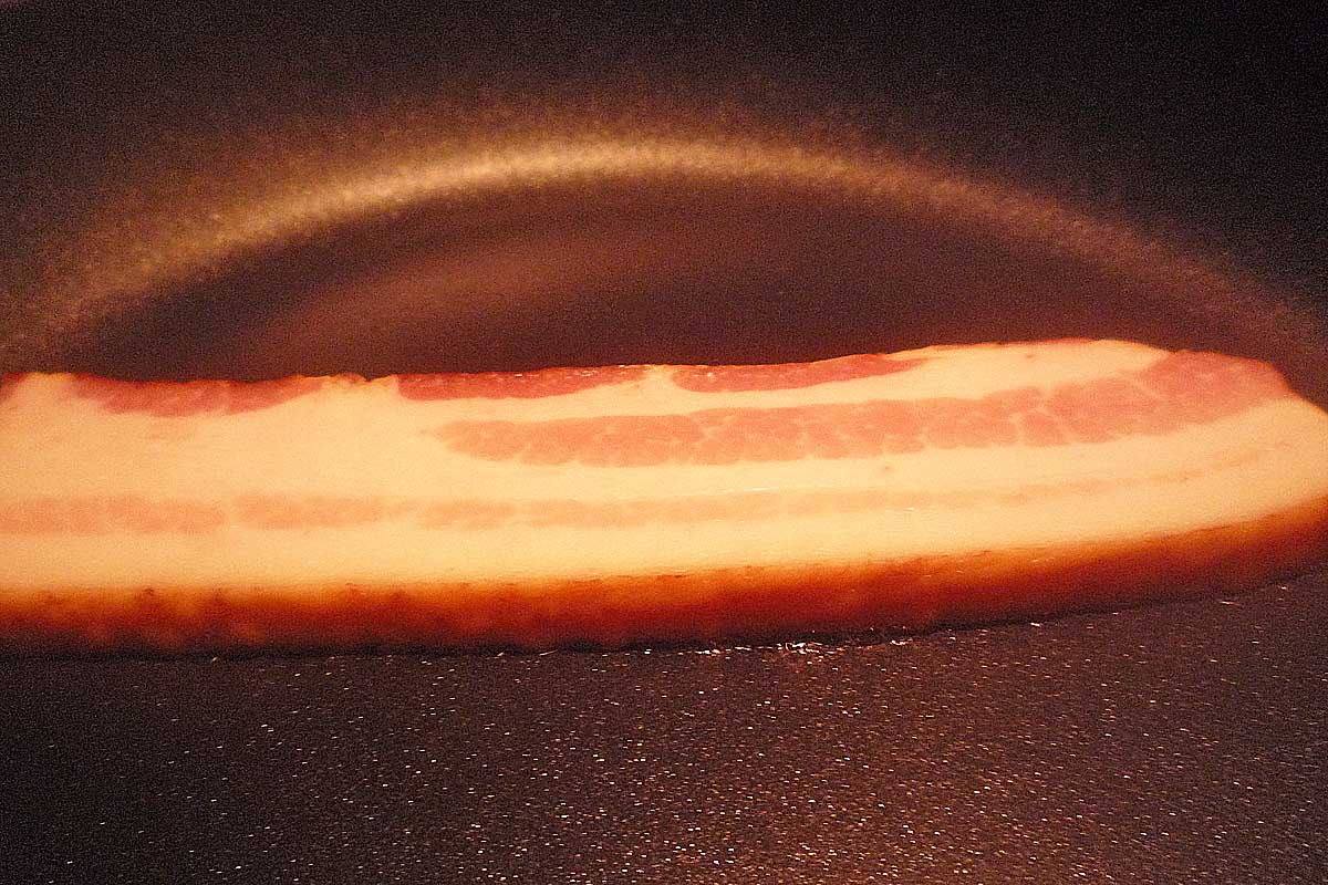 One thick slice of bacon in a black pan, just beginning to cook.
