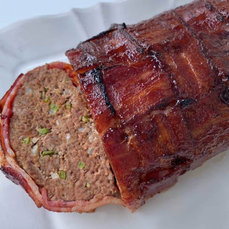 close up photo of bacon weave meatloaf on a white platter, with one sliice cut on the left side
