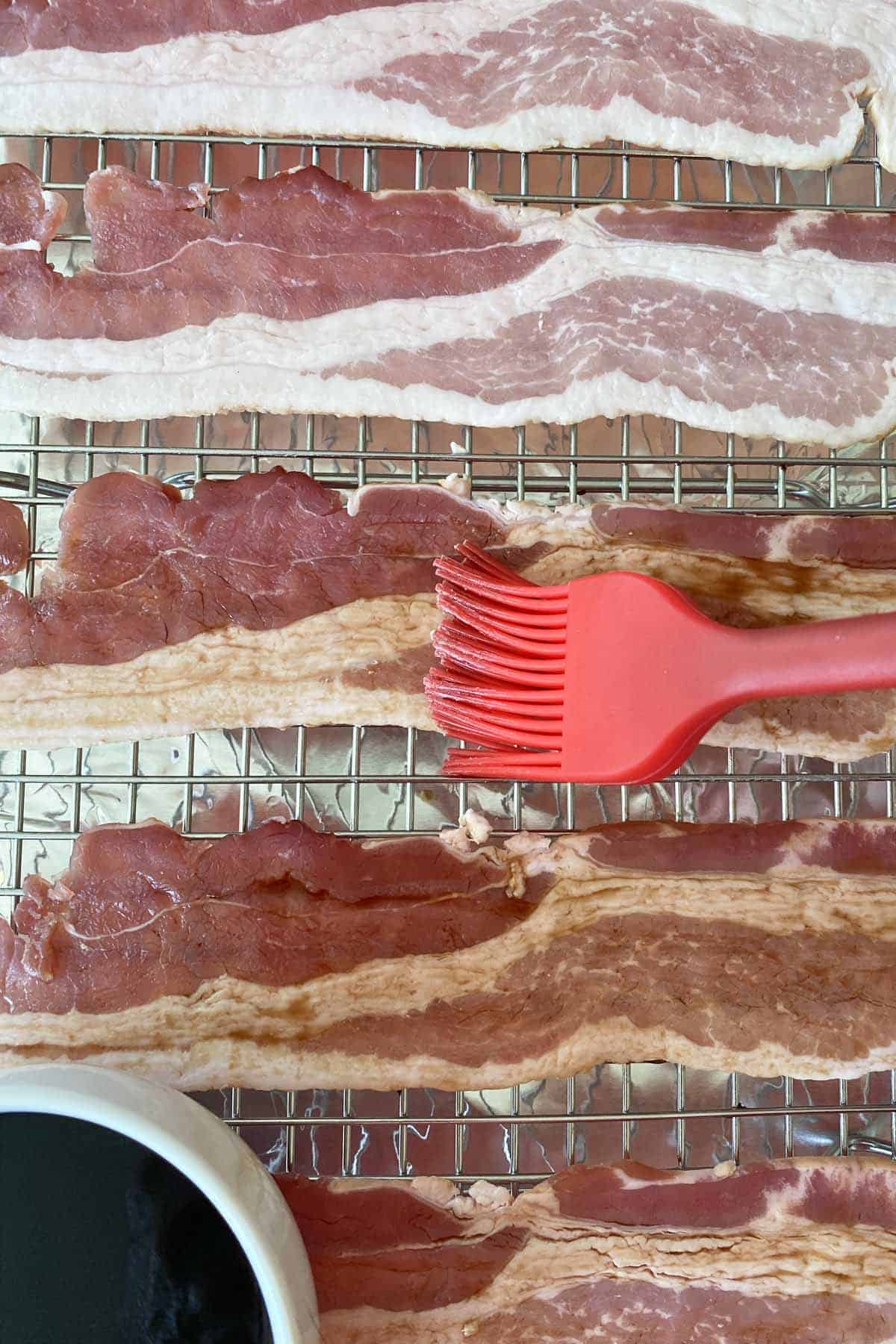 5 slices of bacon on a wire baking rack being brushed with Liquid Smoke with a red silicone brush.