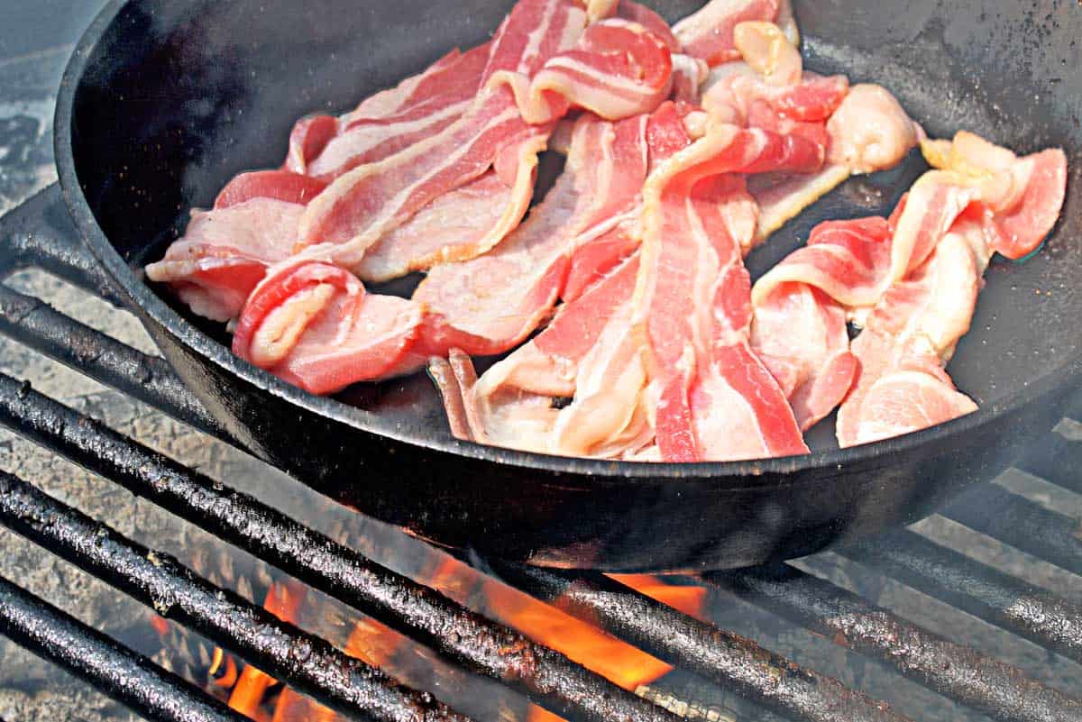A pound of bacon slices cooking in a large cast iron skillet over a grill grate with fire beneath.