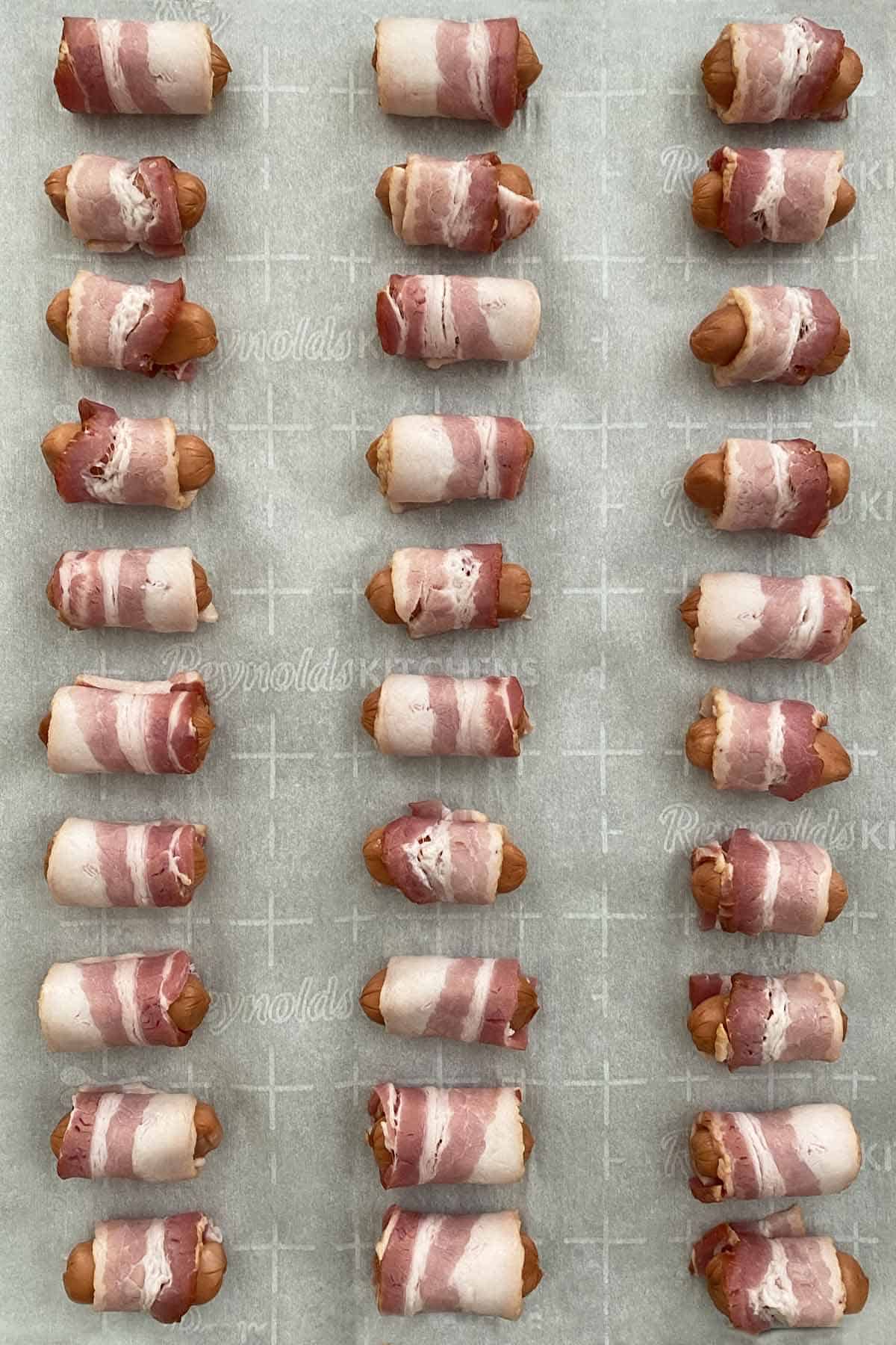 A parchment paper lined baking sheet arranged with 30 uncooked bacon wrapped lil smokies sausages