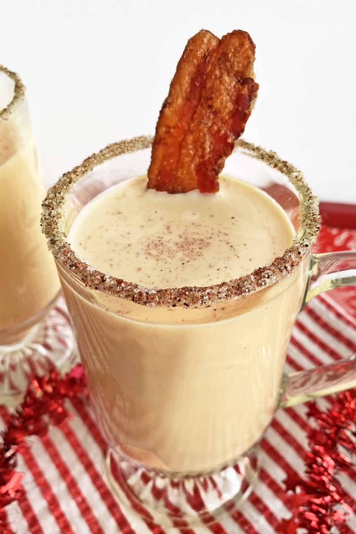 A glass mug filled with bourbon eggnog and topped with a strip of candied bacon, on a red and white napkin.