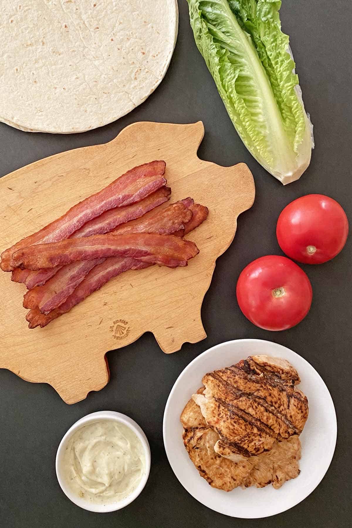 Flour tortillas, a head of romaine lettuce, 6 cooked bacon strips, 2 tomatoes, ranch mayonnaise, and 2 grilled chicken breasts.