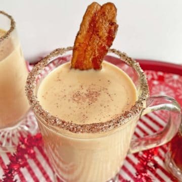 A glass mug of bourbon eggnog topped with a slice of maple candied bacon, on a red and white napkin,