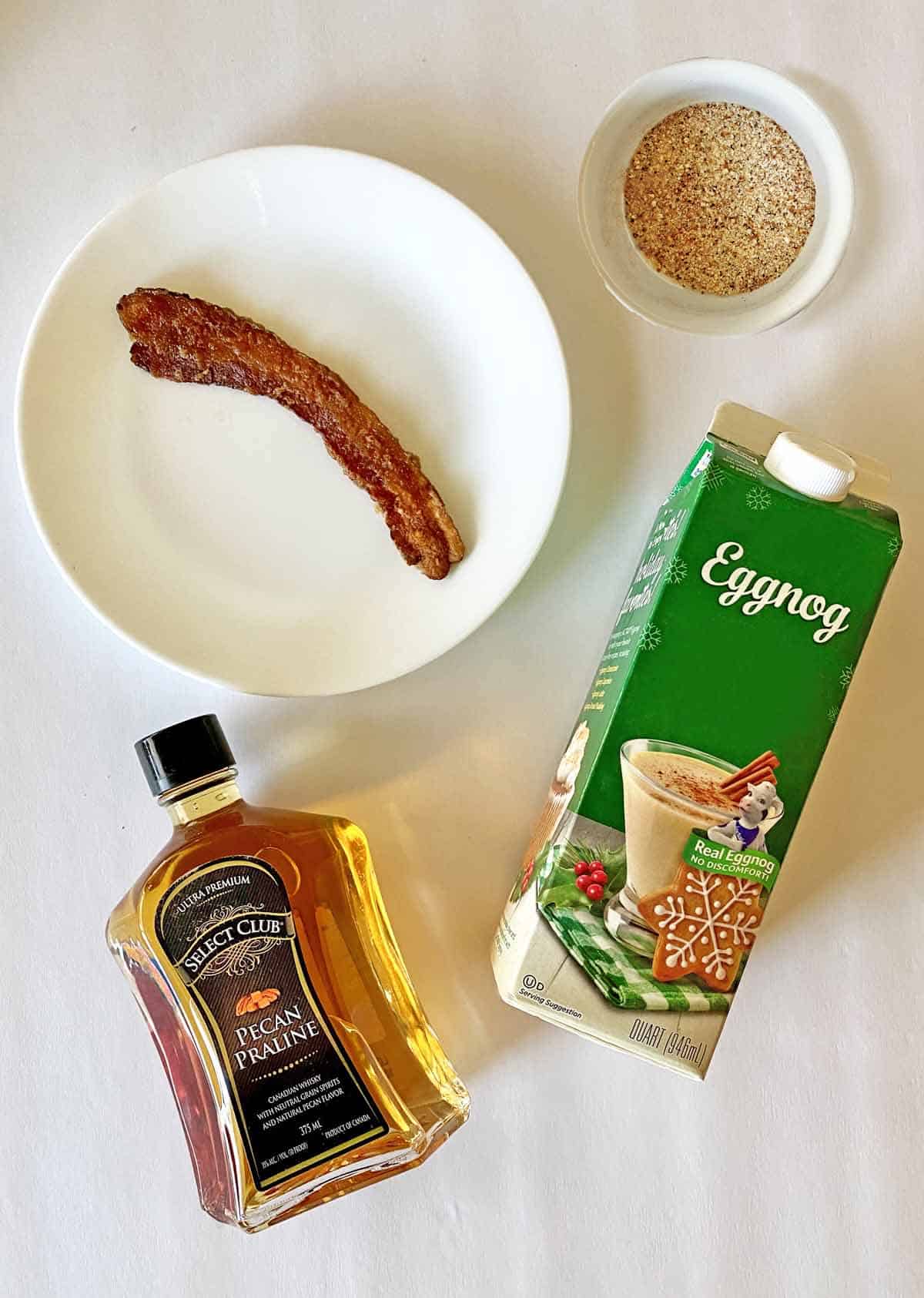 From top, a dish with nutmeg and sugar, a strip of candied bacon, a carton of eggnog and a bottle of pecan praline bourbon whiskey.