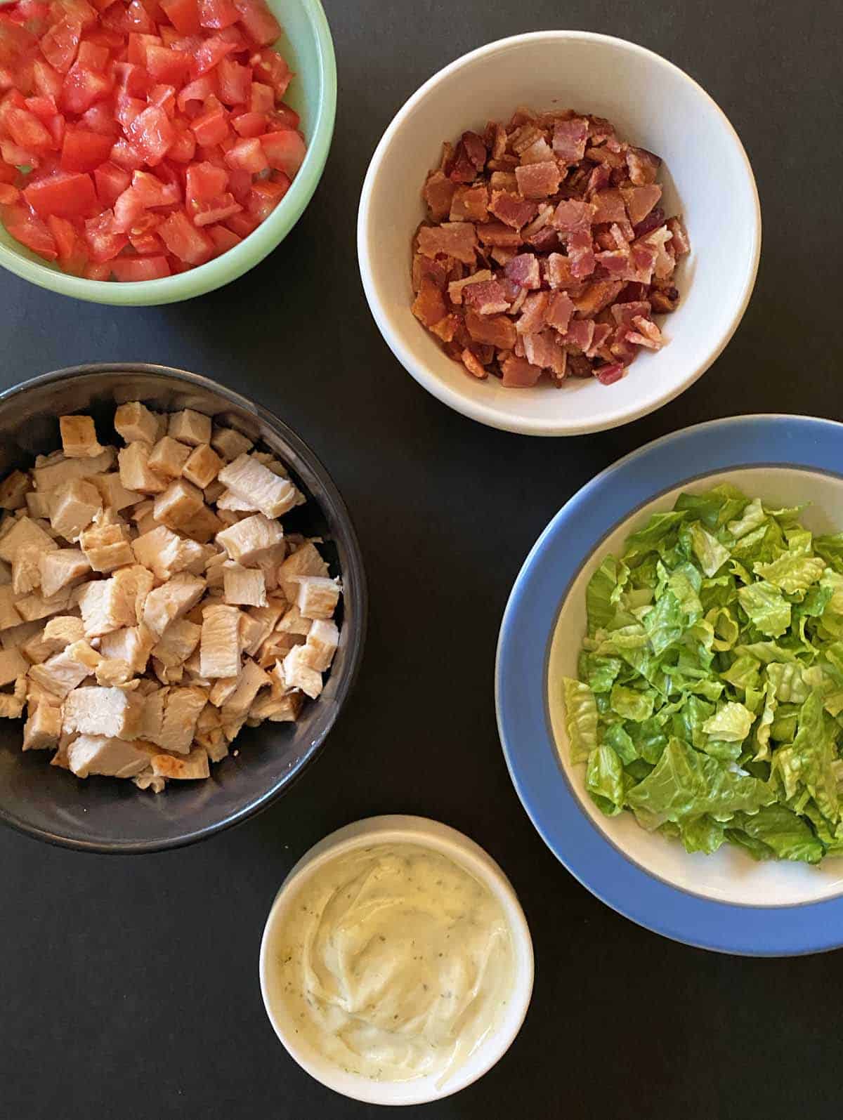 Small bowls containing chopped tomato, bacon, chicken, lettuce and ranch mayo. 