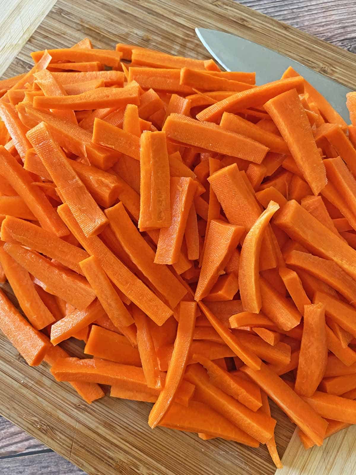 A pile of peeled, cut carrot sticks on a wood cutting board with a knife laid beside it.