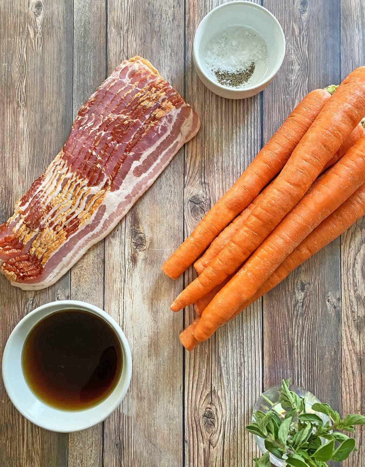 A slab of sliced bacon, six large carrots, a small bowl of maple syrup, and a glass vase with fresh oregano sprigs.