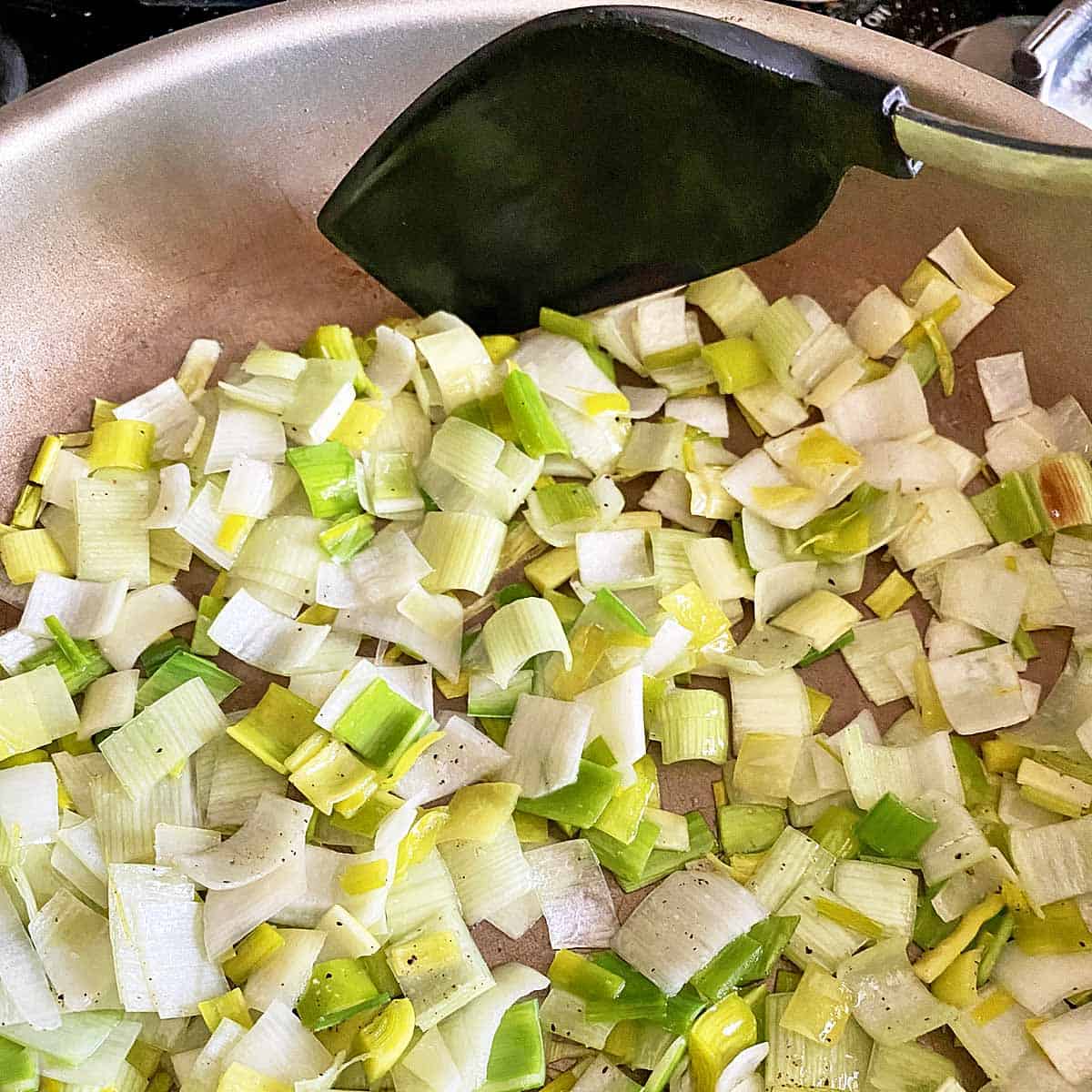 Chopped leeks cooking in a skillet with a black spoon stirring them.