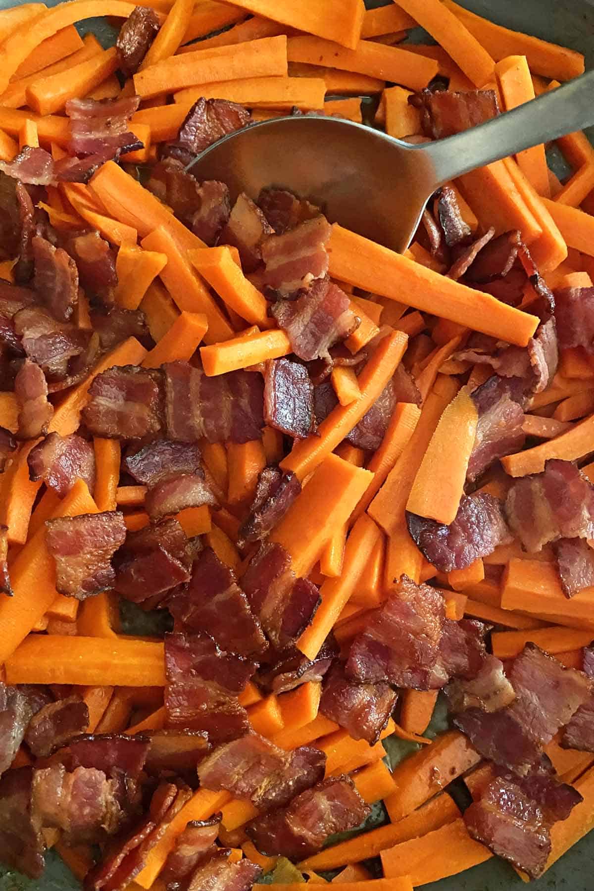 Close up photo of a silver spoon stirring cooked bacon and carrots in a skillet.