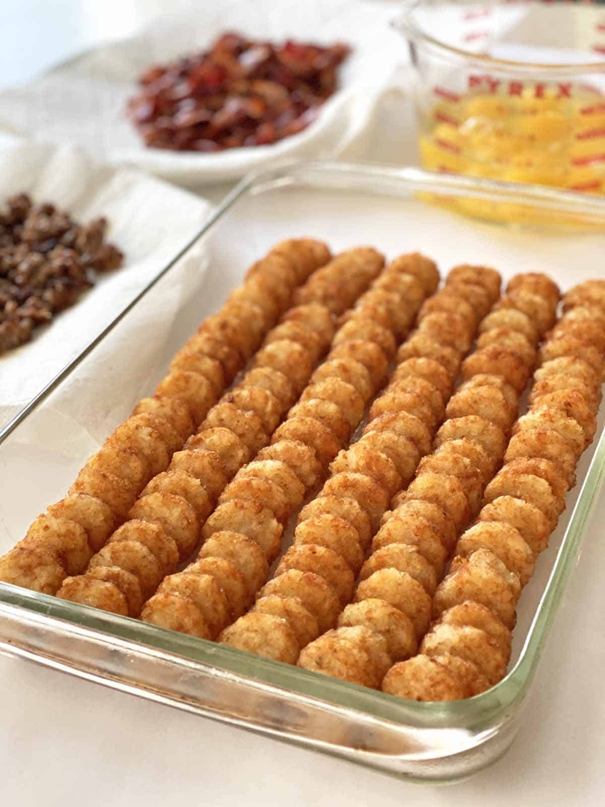 Tater tots arranged in a glass baking dish, with plates of cooked sausage and bacon and a measuring cup of eggs in the background.