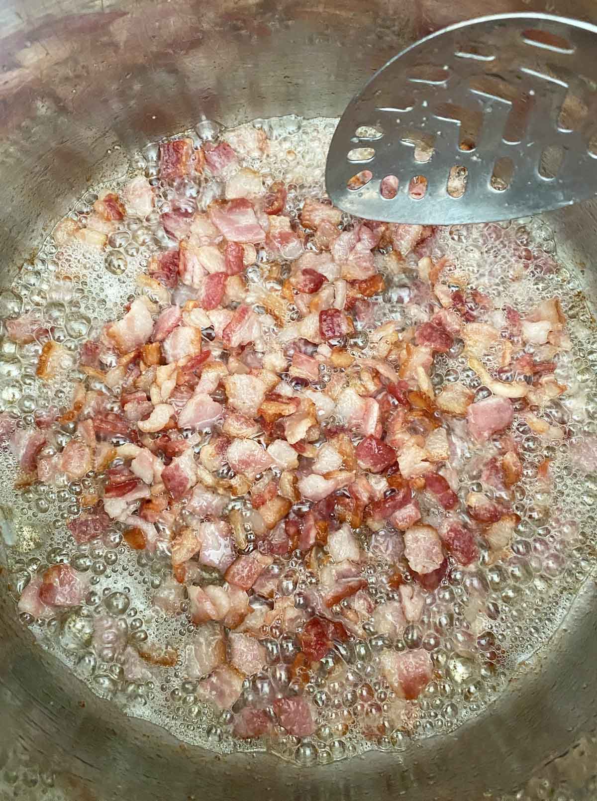 Chopped bacon cooking in a pot with a silver slotted spoon stirring.