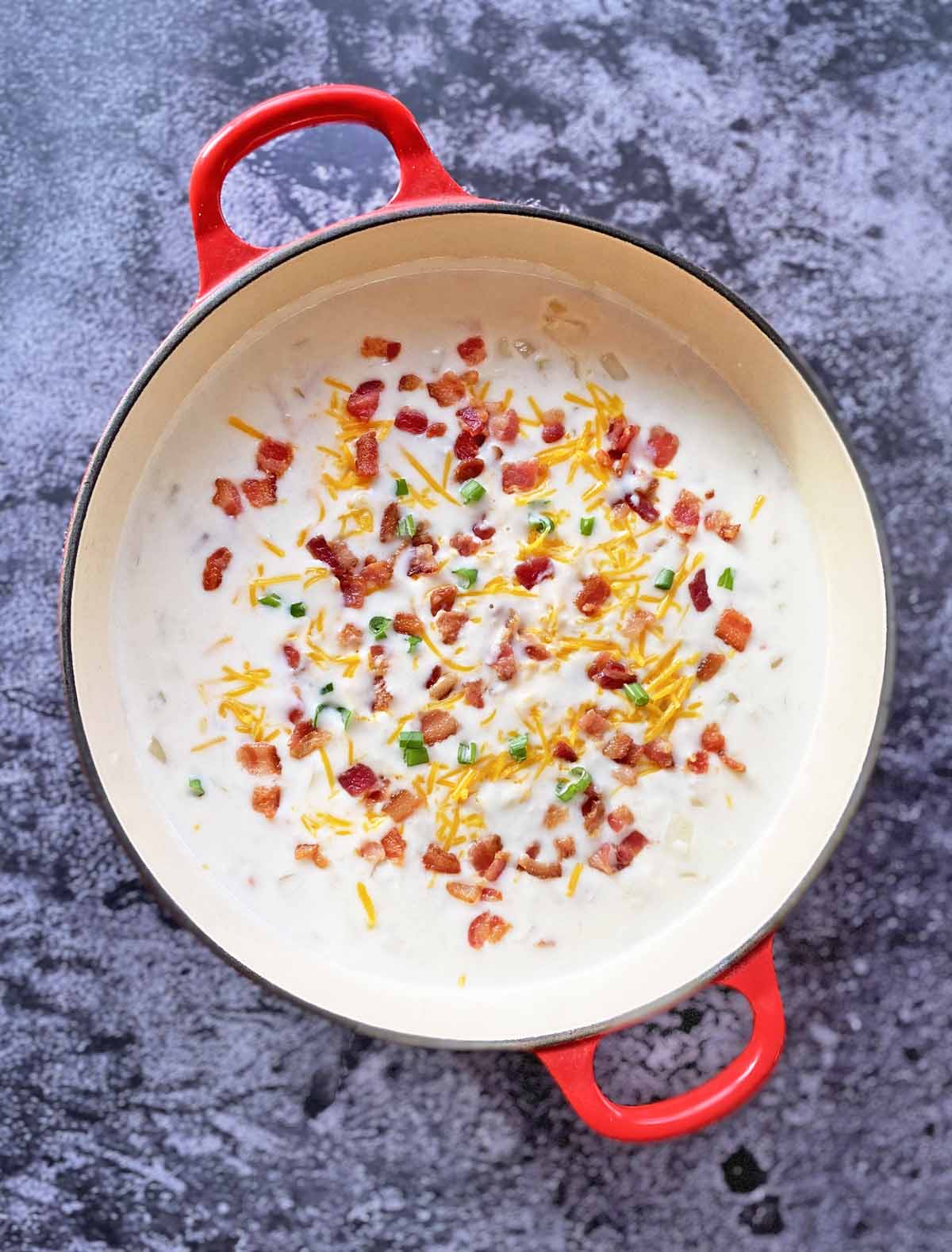 A red handled pot filled with cream of bacon soup garnished with cheese and green onions.