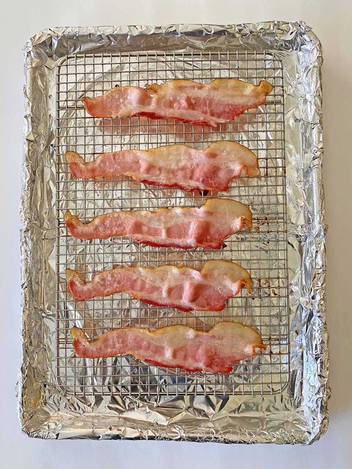 Six slices of partially cooked bacon on a wire rack atop a foil-lined baking sheet. 