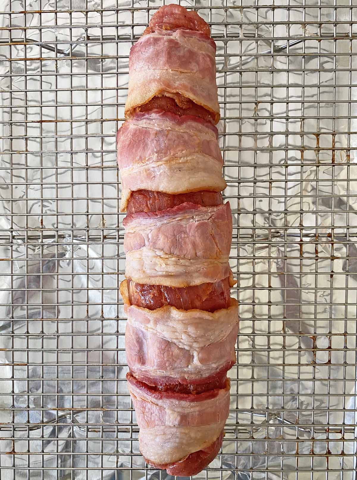 Uncooked pork tenderloin wrapped with strips of partially cooked bacon on a wire baking rack.