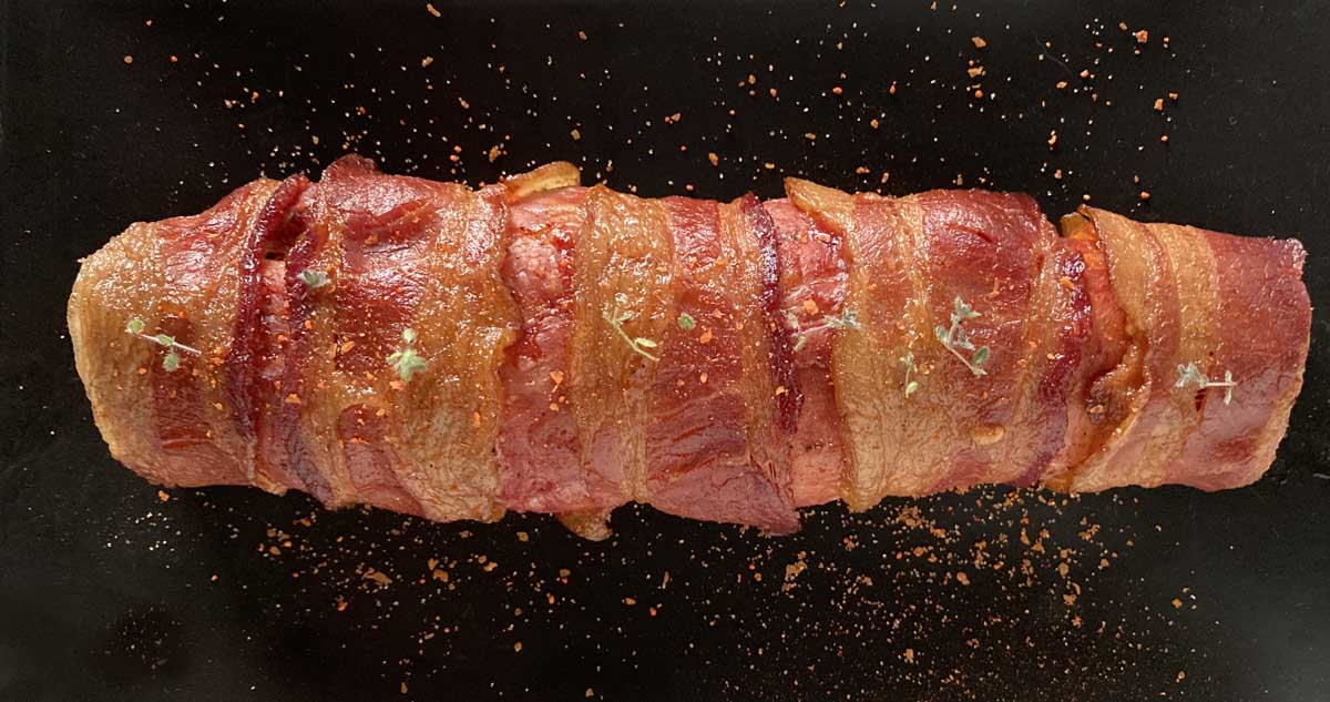 A whole bacon wrapped smoked pork tenderloinl on a black platter.
