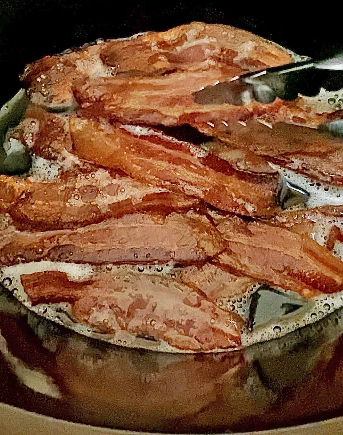 Cooked bacon inside the crock pot, being rearranged with silver tongs.
